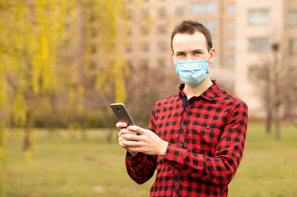 Kaukaski mężczyzna noszący koszulę i maskę ochronną przed wirusem stojącym na zewnątrz przy użyciu smartfona komórkowego do wysyłania wiadomości przeglądających rozmowy wideo przez internet - Zdjęcie, obraz