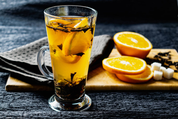 Citrus drink, tea with petals in a transparent glass mug with slices of sweet orange and refined sugar. Slicing fruit on a wooden Board with a brown napkin in a cozy dark background in a low key - Foto, immagini