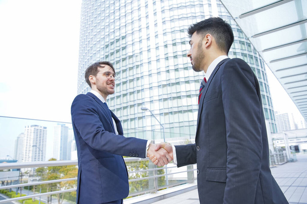 Dois homens de negócios apertando as mãos com um sorriso no distrito de negócios
 - Foto, Imagem