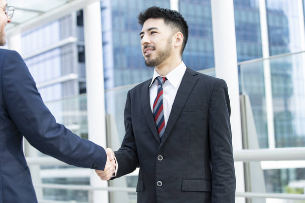 Dos hombres de negocios estrechando la mano con una sonrisa en el distrito de negocios
 - Foto, Imagen
