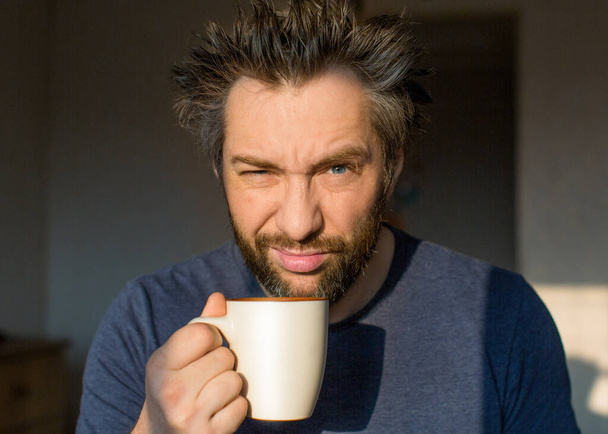 Смешной человек с бородой после пробуждения с большой чашкой кофе или чая смотрит в камеру одним глазом
. - Фото, изображение