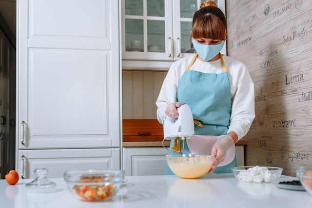 A woman cook in a medical mask and gloves whips up dough for an Easter cake - Fotoğraf, Görsel