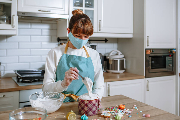 A cook in a medical mask and gloves in a home kitchen puts sweet figures on the icing of an Easter cake - Photo, image