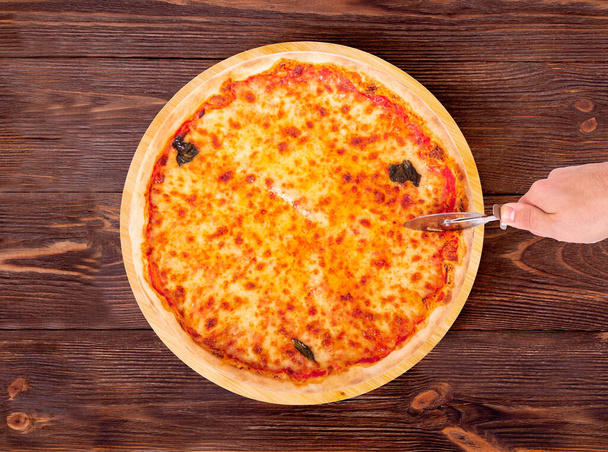Miehen käsi leikkaa margherita-pizzan pizzaleikkurilla. Herkullinen pizza mozzarellalla, bocconcinilla ja basilikan lehdillä pyöreällä puuvadilla, joka on puisella maalaispöydällä
 - Valokuva, kuva