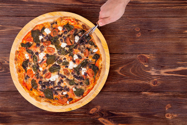 Miehen käsi leikkaa pizzan pizzaleikkurilla, ylhäältä ja kopioi tilaa. Herkullinen pizza kirsikkatomaateilla, pinaatilla, mozzarellalla, fetalla, kalamata-oliiveilla ja sienillä pyöreällä puulevyllä, joka on puisella maalaispöydällä.
 - Valokuva, kuva