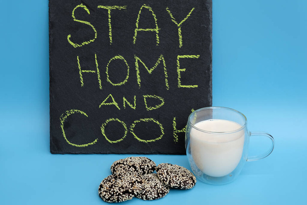 Glass of milk and chocolate chip cookies on a blue background. The inscription on the black chalkboard stay home and cook. The concept -quarantine, what to do in isolation at home during the epidemic - Photo, image