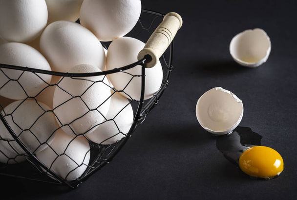 Kuřecí vejce v kovovém koši na černém dřevěném stole. Prasklé vejce přímo na stole se žlutým žloutkem a rozbitou skořápkou. Koncept obrazu pro přísloví: nedávejte všechna vajíčka do jednoho košíku - Fotografie, Obrázek