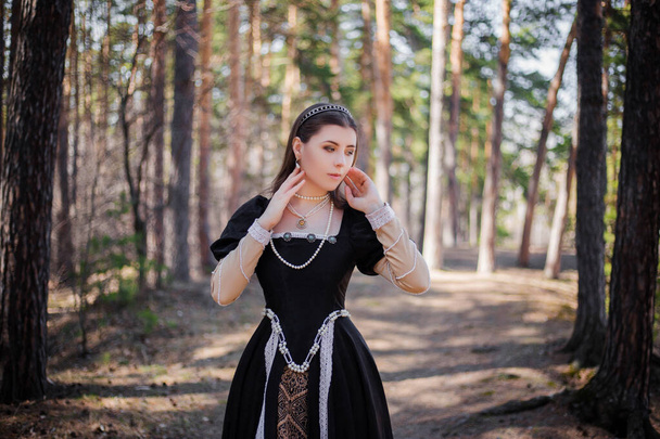 Portrait of a young, beautiful woman in a black medieval dress standing in a pine forest. Attractive girl in a gothic dress. - Foto, Bild