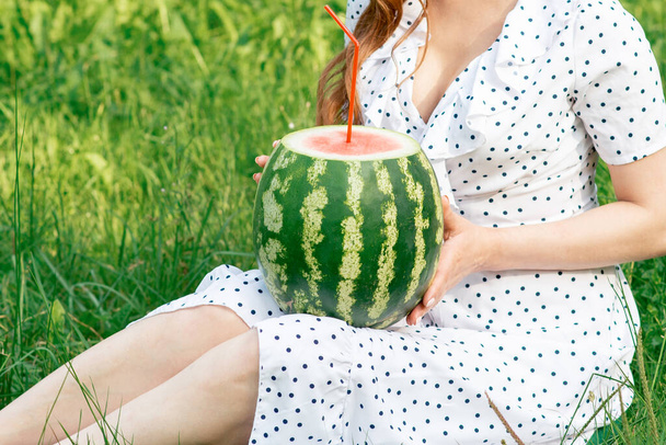 Girl holds whole watermelon with cocktail straw on green grass background. - Fotografie, Obrázek