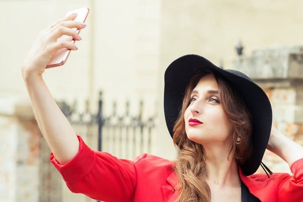 Beautiful girl in black hat makes selfie herself at city street. - Photo, Image