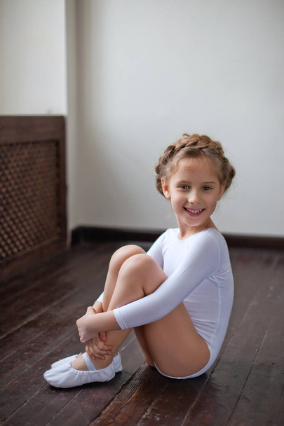Urocza dziewczynka w białym stroju gimnastycznym trenuje gimnastykę rytmiczną. Mała dziewczynka ćwiczy na podłodze. Dla dziecka, koncepcja - Zdjęcie, obraz