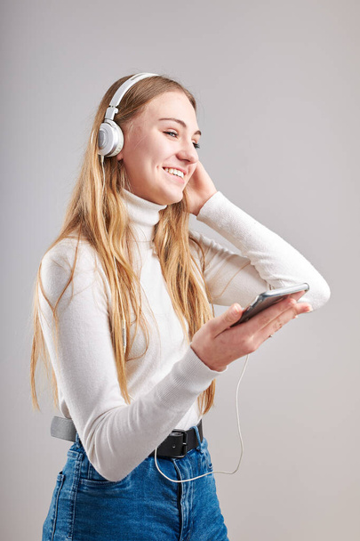 Young woman girl listening to music streaming content having fun watching video enjoying video chat talking with friends making gestures faces using smartphone earphones headphones standing over plain grey background - Fotó, kép
