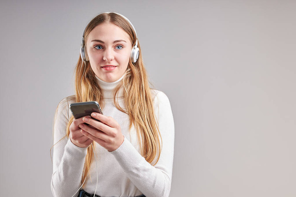 Young woman girl listening to music streaming content having fun watching video enjoying video chat talking with friends making gestures faces using smartphone earphones headphones standing over plain grey background - Φωτογραφία, εικόνα
