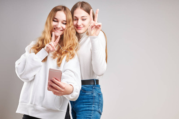 Young women girls listening to music together streaming content having fun watching video enjoying video chat talking with friends making gestures faces using smartphone earphones headphones standing over plain grey background - Zdjęcie, obraz