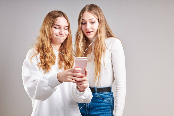 Young women girls listening to music together streaming content having fun watching video enjoying video chat talking with friends making gestures faces using smartphone earphones headphones standing over plain grey background - Valokuva, kuva