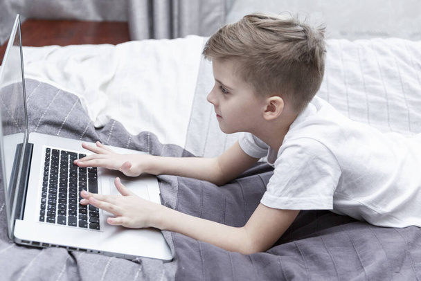 A boy of 8 years old is lying on the bed and uses a laptop. Distance learning during the coronavirus pandemic. Close-up. - Foto, imagen