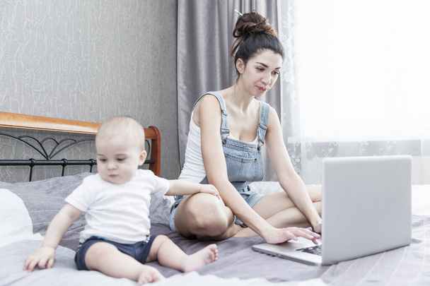 Trabajando en casa mamá con el bebé están sentados en la cama. Internet y trabajo remoto
. - Foto, imagen
