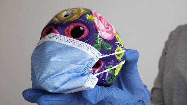 Χέρια από καουτσούκ ιατρικά γάντια που κρατούν διακοσμημένα χρωματιστά κρανία φορώντας μια μπλε χειρουργική μάσκα. - Φωτογραφία, εικόνα