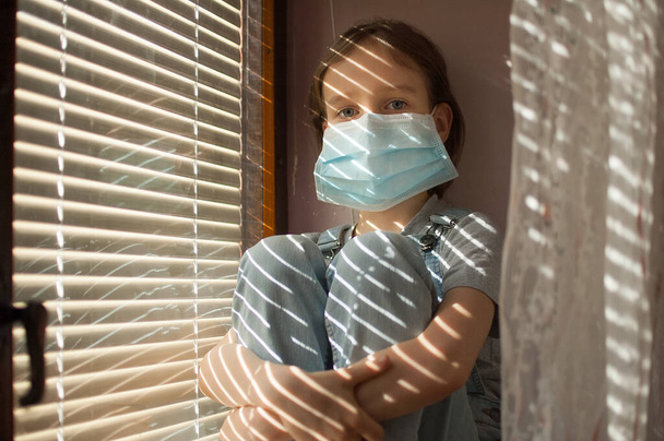 Tired bored little girl is sitting on the windowsill in protective disposable mask on her face during self-isolation at home because of Coronavirus Covid-19 on blinds background. - Photo, Image