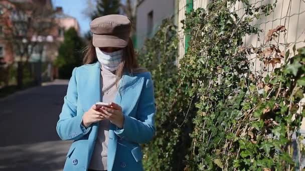 A nő újra felhasználható maszkot visel a szabadban a Coronavirus covid-19 világjárvány idején. Egy lány okostelefont használ az üres utcán. Vigyázz magadra! Tavaszi divat - Felvétel, videó