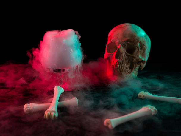 Череп человека возле стекла с густым дымом и костями для Дня мертвых на темном фоне с красным светом
. - Фото, изображение