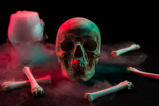Human skull near glass with thick smoke and bones for Day of the Dead on dark background with red and green light. - Foto, afbeelding