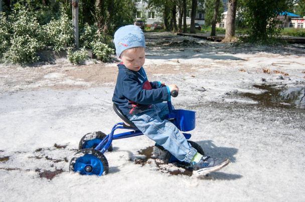Rússia, Novodvinsk - julho de 2019. Um menino loiro de cabelos azuis monta um pequeno triciclo em uma poça completamente coberta com o velho cotão fofo
 - Foto, Imagem