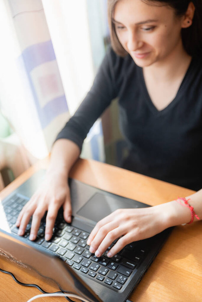Fiatal fehér lány fekete pulóverben nézi a számítógépet, miközben otthon tanul, az ablak mellett. - Fotó, kép
