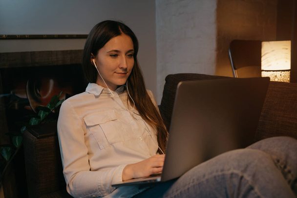 Fernbedienung. Eine kaukasische Frau mit Kopfhörern, die aus der Ferne an ihrem Laptop arbeitet. Eine Geschäftsfrau sitzt auf dem Sofa. - Foto, Bild