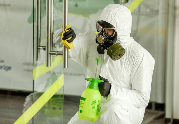A man in protective equipment disinfects with a sprayer in the office. Surface treatment due to coronavirus covid-19 disease. A man in a white suit disinfects the room with a spray gun. Virus pandemic - Photo, image