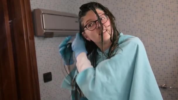 Een meisje verft haar blonde haar in het blauw thuis op de badkamer. Thuis haarverzorging tijdens carantine van coronavirus. - Video