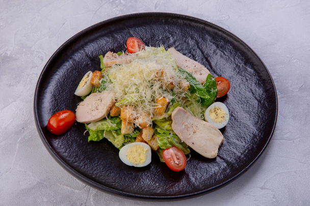 Caesar salad with chicken breast on a dark plate. Home-made chicken fillet, cherry tomatoes, Parmesan cheese, quail eggs, crackers and sauce. European cuisine. Dish from a restaurant or cafe menu - Foto, Imagen