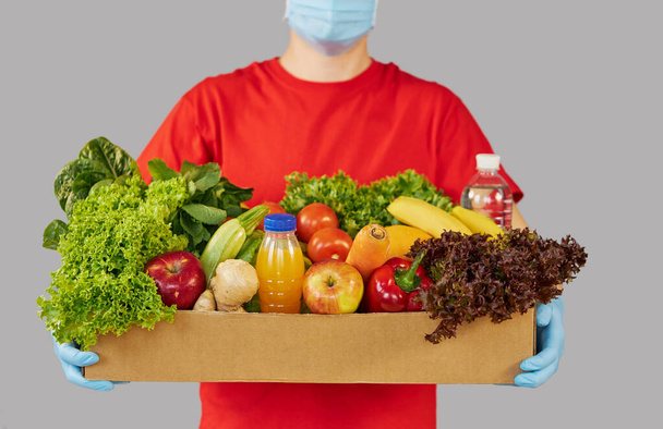 Online shopping concept. Male courier in red uniform, protective mask and gloves with a grocery box with fresh fruits and vegetables. Home delivery food during quarantine coronavirus  - Valokuva, kuva