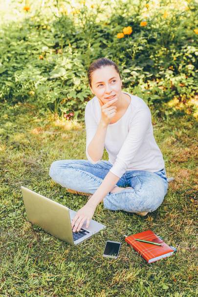 Mobile Office. Freelance business concept. Young woman sitting on green grass lawn in city park working on laptop pc computer. Lifestyle authentic candid student girl studying outdoors - Photo, Image