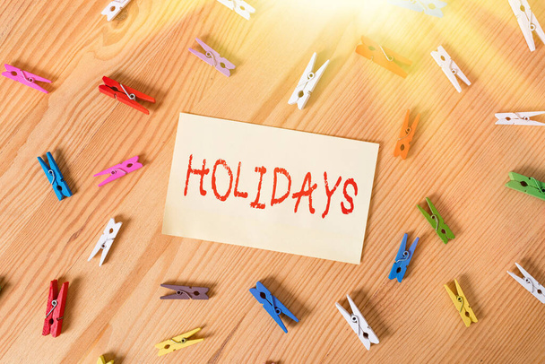 Handwriting text Holidays. Concept meaning an extended period of leisure and recreation spent away from home Colored clothespin papers empty reminder wooden floor background office. - Photo, Image