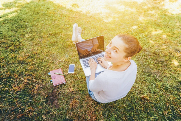 Mobile Office. Freelance business concept. Young woman sitting on green grass lawn in city park working on laptop pc computer. Lifestyle authentic candid student girl studying outdoors - Zdjęcie, obraz