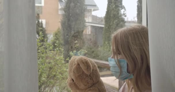 Side view of adult blond Caucasian woman in face mask holding teddy bear as sitting on windowsill. Portrait of adult sad lady looking out at snow outdoors. Isolation, lockdown. Cinema 4k ProRes HQ. - Materiał filmowy, wideo