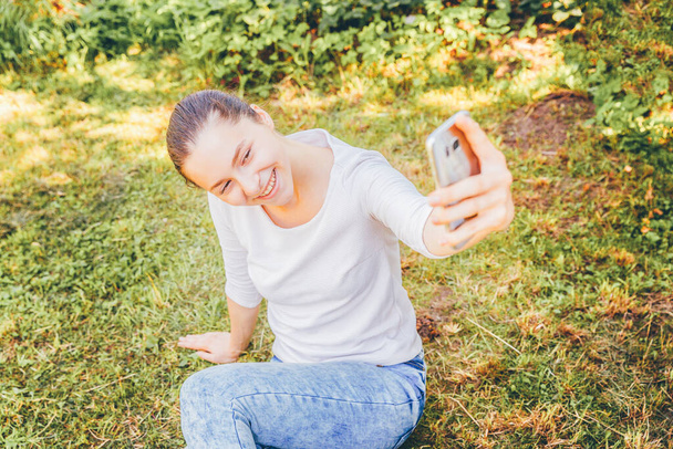 Young funny girl take selfie from hands with phone sitting on green grass park or garden background. Portrait of young attractive woman making selfie photo on smartphone in summer day - Fotoğraf, Görsel