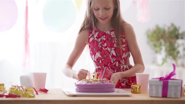 Caucasian girl is dreamily smiling and looking at birthday rainbow cake. Festive colorful background with balloons. Birthday party and wishes concept. - Footage, Video