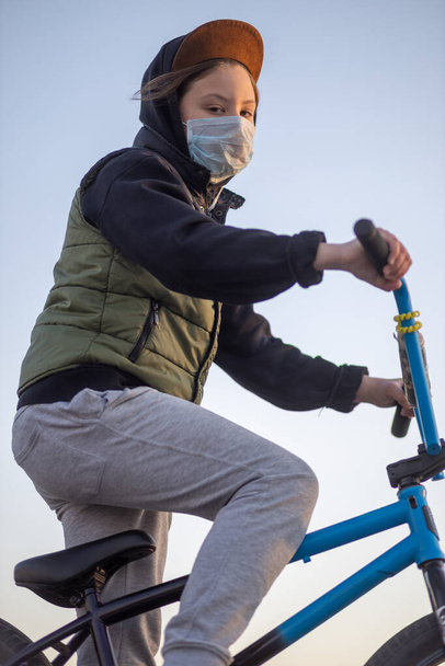 Egy tinédzser portréja a naplementében, álarcban egy kék biciklin. Karantén. Biciklizni tilos. Kovid-19. - Fotó, kép