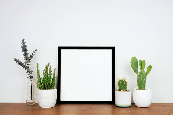 Mock up schwarzen quadratischen Rahmen mit Topfpflanzen und Zweigdekor. Holzregal vor einer weißen Wand. Kopierraum. - Foto, Bild