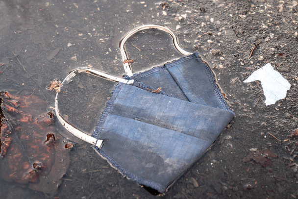 A dirty used homemade blue cloth face mask covering or medical mask with elastic straps and thread lays littering the road in a puddle during the COVID-19 pandemic and outbreak. - Photo, Image