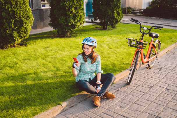 Soggetto ecologico modo di trasporto bicicletta. Bella giovane donna caucasica che indossa un casco blu e pose di capelli lunghi in piedi accanto a una bici a noleggio di colore arancione con un cesto in un parco cittadino. - Foto, immagini