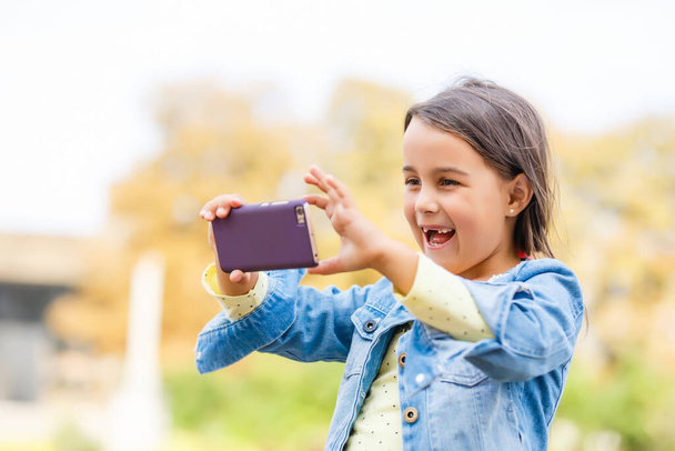 ビデオストリーミング。愛らしい子供は新しい技術を学ぶ。携帯電話を使う少女。スマートフォンを持つ小さな女の子の子供。かわいい携帯電話技術のユーザー。モバイルデバイスで動画を見る. - 写真・画像