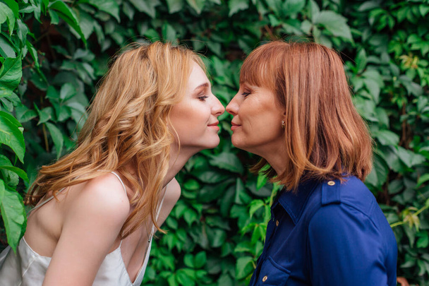 Beautiful mother and her grown up daughter standing nose to nose to each other on a summer day outside on a green nature background - Zdjęcie, obraz