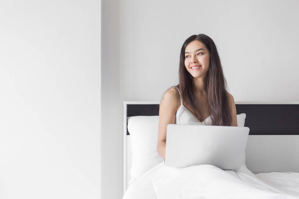 若いです幸せな美しさアジアの女性座っているベッドの上にコンピュータのラップトップ. - 写真・画像