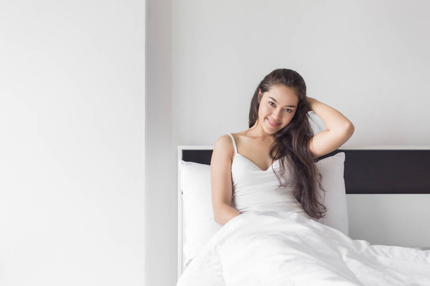 Νεαρή ευτυχισμένη ομορφιά Ασιάτισσα γυναίκα κάθεται στο κρεβάτι με χαμογελαστό πρόσωπο. - Φωτογραφία, εικόνα