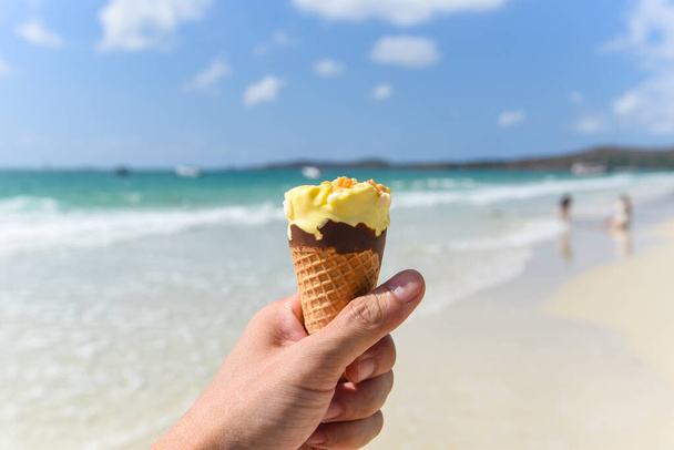 Melting ice cream on beach in summer hot weather ocean landscape nature outdoor vacation , Yellow ice cream mango with nuts / Ice cream cone in hand with sea background - Photo, Image