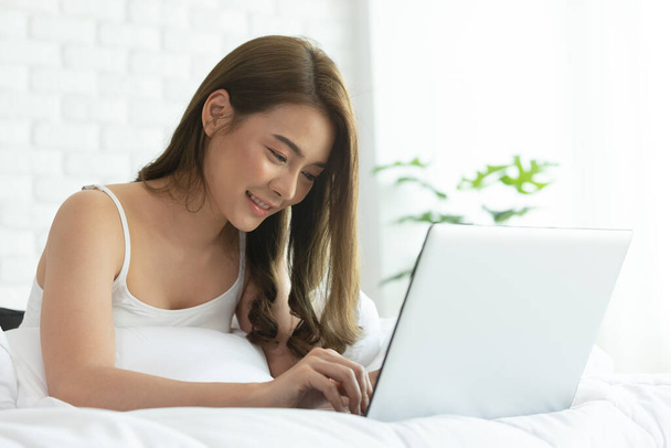 Красива азіатська жінка працює на ноутбуці, сидячи на ліжку в будинку. Жінка перевіряє соціальні додатки і працює. Робота з домашньої концепції
. - Фото, зображення