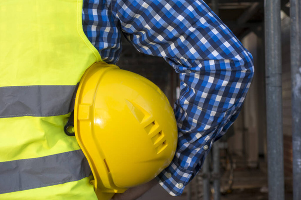 Immagine ritagliata di un operaio edile che indossa un giubbotto di sicurezza giallo e tiene un casco giallo sotto le ascelle
 - Foto, immagini